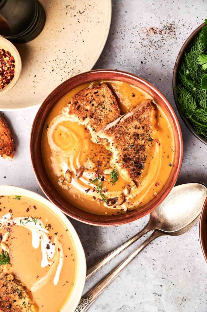 A bowl of creamy butternut squash soup garnished with herbs, nuts, and two pieces of toasted bread, placed on a grey tabletop alongside a spoon and a bowl of fresh herbs.