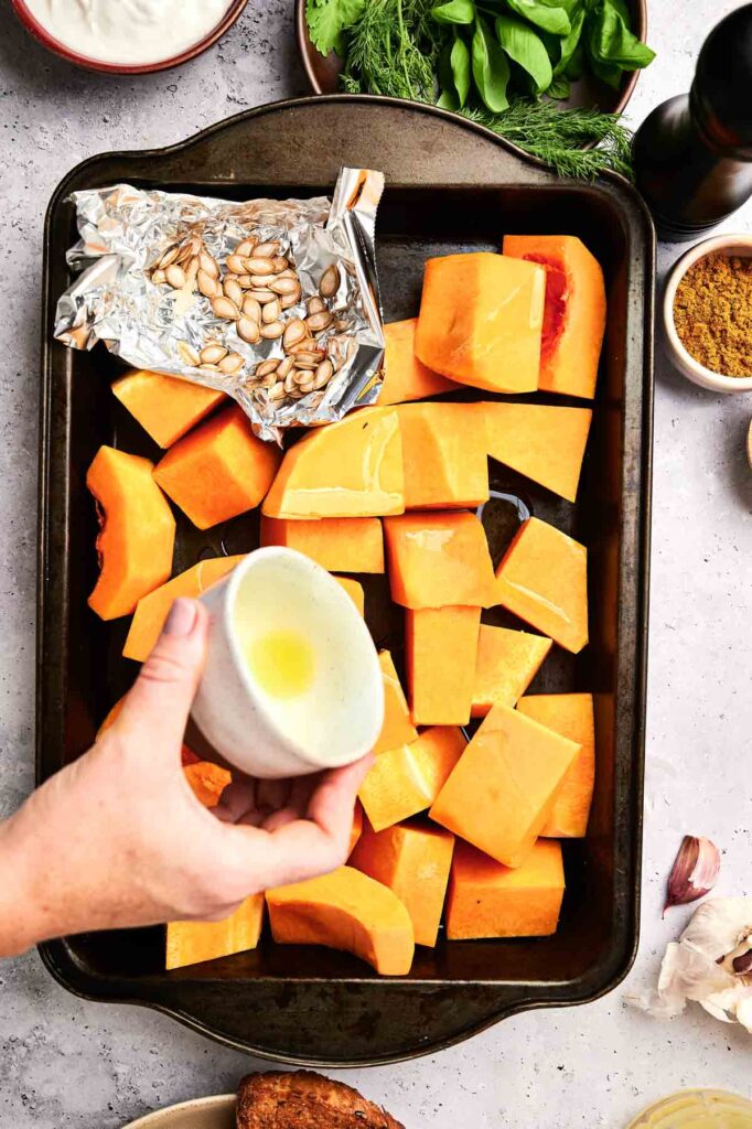 A hand pours oil from a small cup over cubed butternut squash and seeds on a baking tray, surrounded by various ingredients—perfect preparation for a rich and flavorful Butternut Squash Soup.