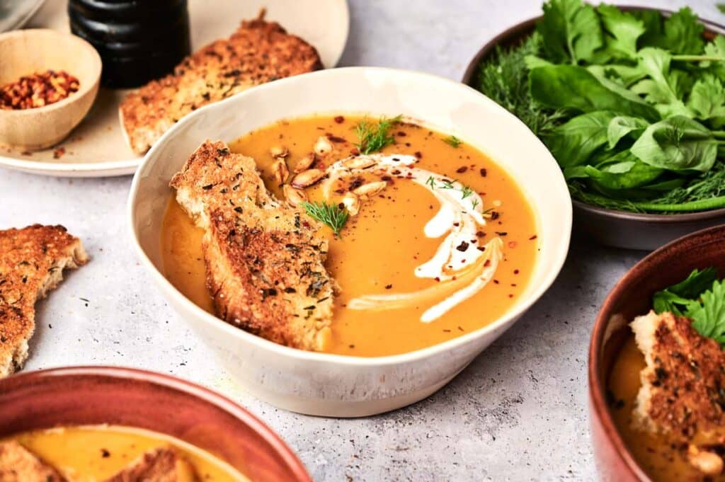 A bowl of creamy Butternut Squash Soup garnished with a drizzle of cream, a piece of bread, and herbs. Plates with more bread, herbs, and spices are on the table.