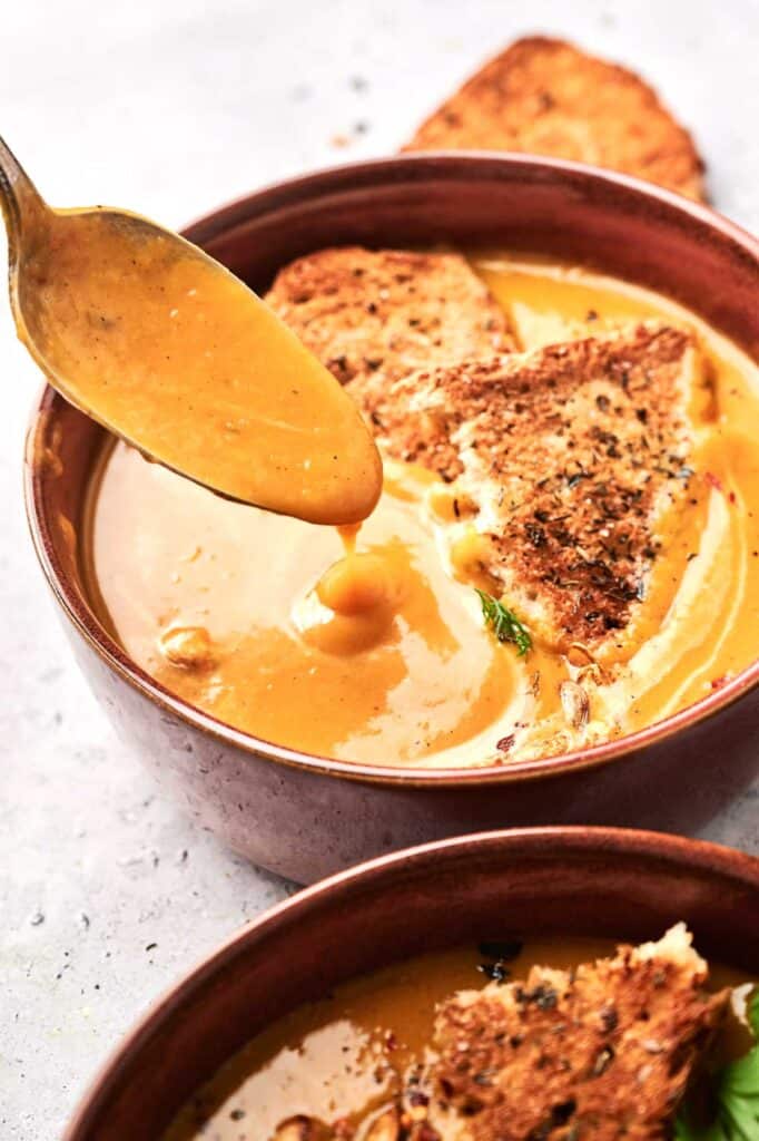 A spoonful of creamy Butternut Squash Soup being served into a brown bowl with toasted bread slices.