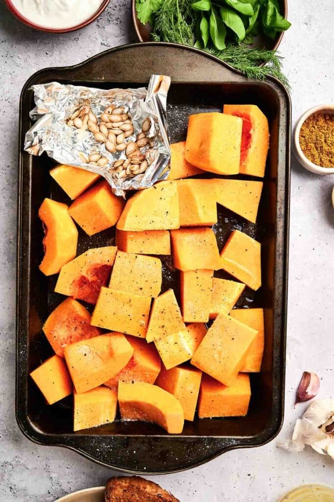 A baking sheet filled with cubed butternut squash, seasoned with salt and pepper, ready to transform into a delicious Butternut Squash Soup. A small piece of foil holds butternut squash seeds. Various ingredients surround the sheet.