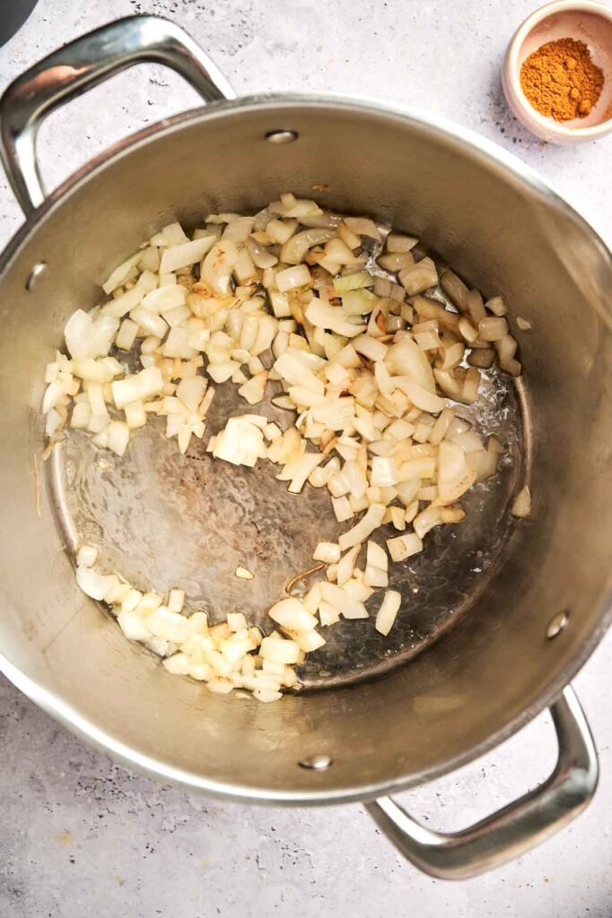A pot with chopped onions being saut&eacute;ed on a stovetop, the first step in preparing a hearty Butternut Squash Soup. A small bowl containing a powdered spice sits beside the pot.