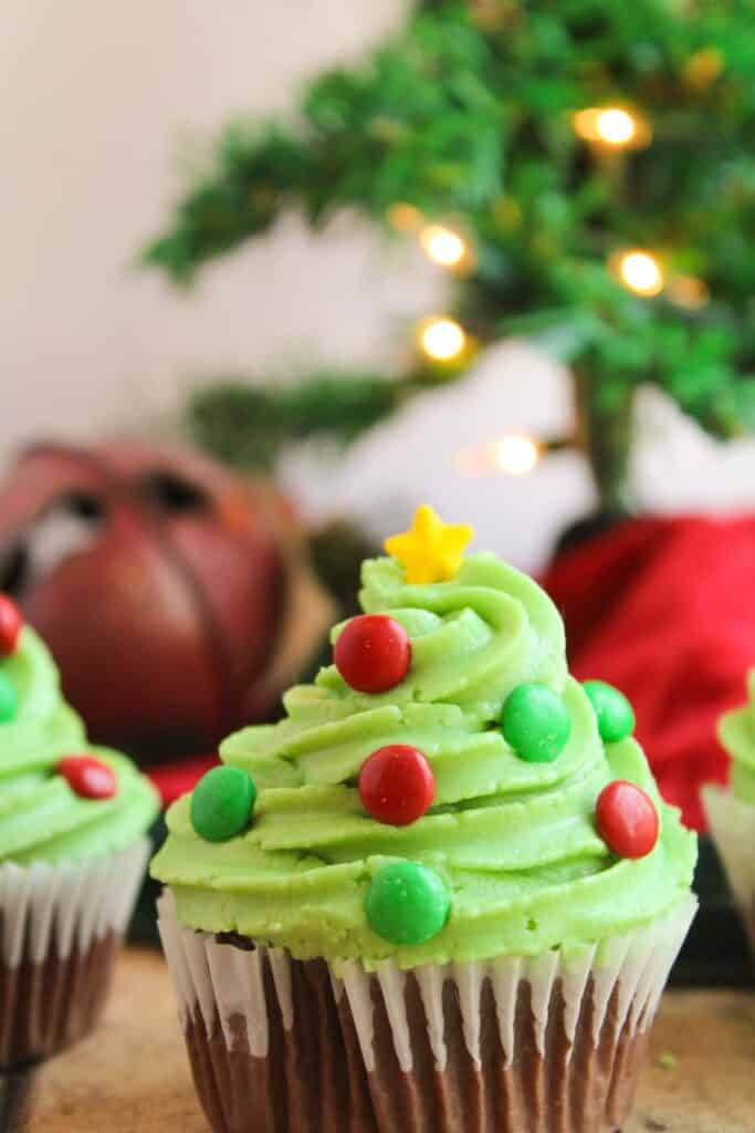 A festive Christmas tree cupcake is decorated with green frosting to resemble a Christmas tree, garnished with red and green candies, and topped with a star. In the background, you can see a blurred Christmas tree.