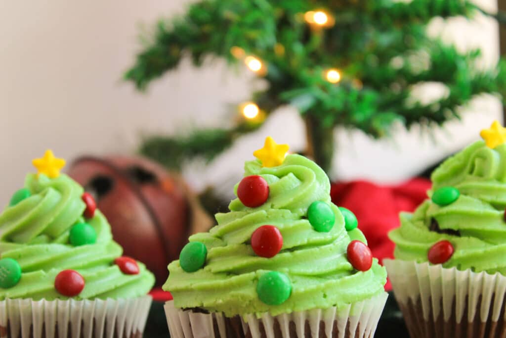 Three festive Christmas tree-shaped cupcakes with green frosting, adorned with red and green candies and a yellow star topper, sit proudly in front of a blurred Christmas tree.