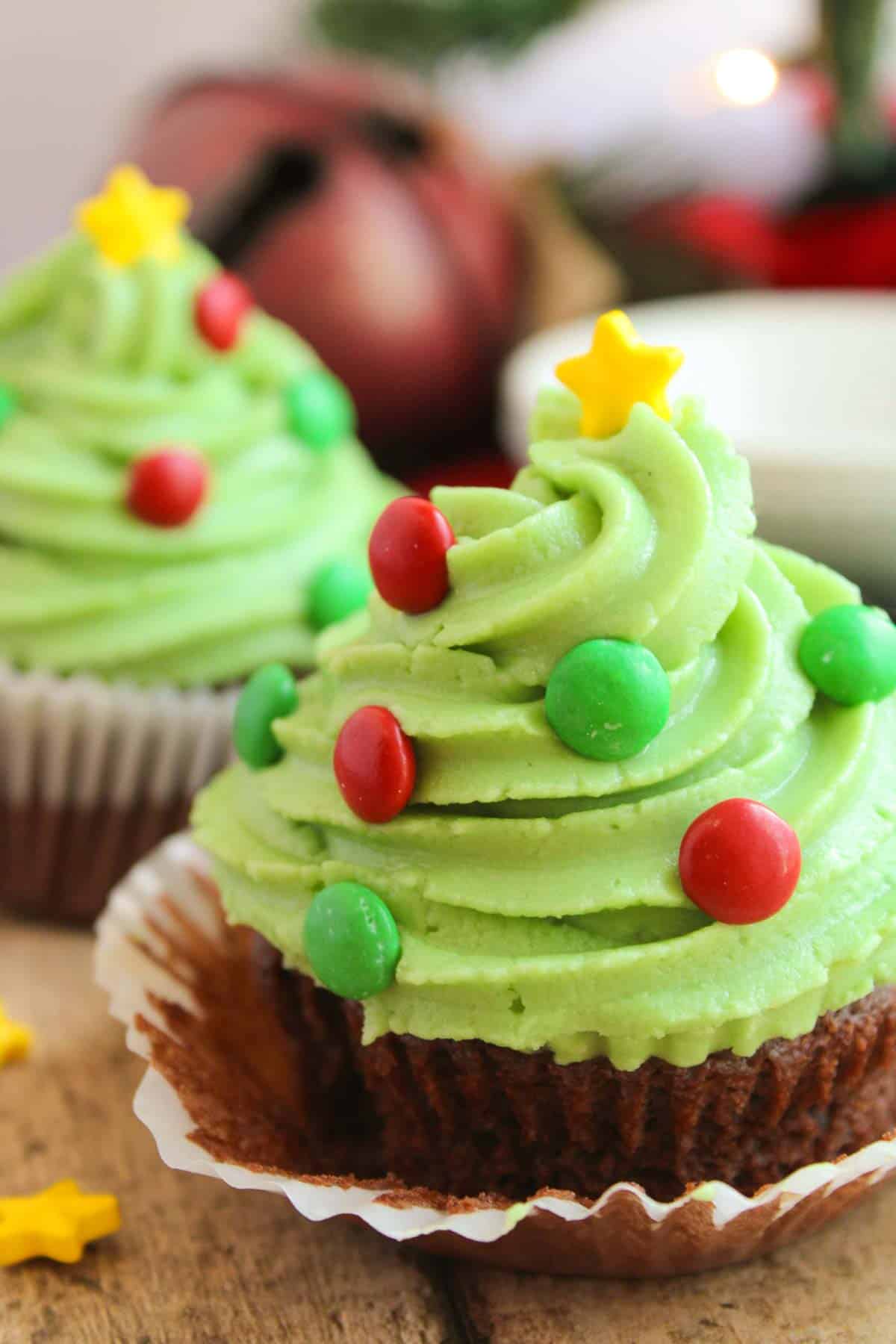 Two Festive Christmas Tree Cupcakes with green frosting shaped like Christmas trees, decorated with red and green candy ornaments and yellow star toppers, sit on a wooden surface.