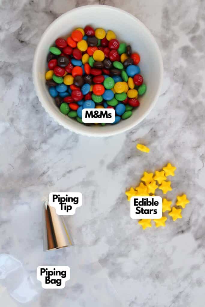 A white bowl filled with M&Ms, a small pile of yellow edible star shapes, a piping bag, and a piping tip are arranged on a marble surface, ready to decorate Festive Christmas Tree Cupcakes.