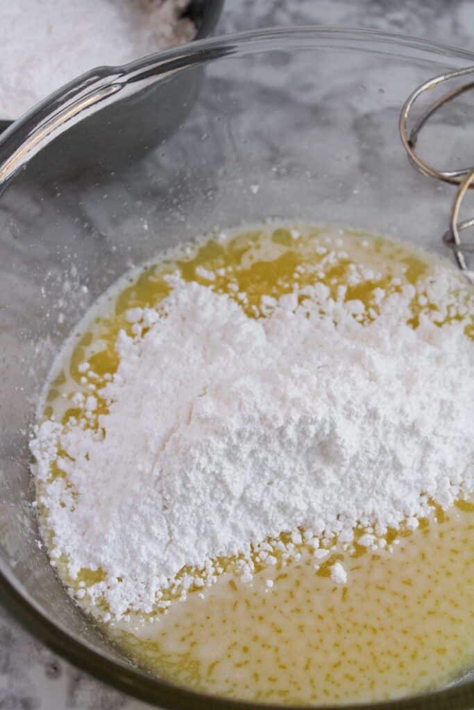 A glass bowl contains a mixture of melted butter and powdered sugar, with a whisk partially visible on the right, ready to be transformed into creamy frosting for festive Christmas Tree Cupcakes.
