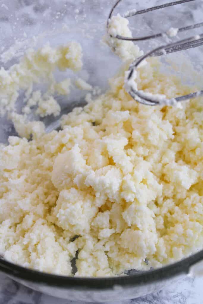 A close-up of clumped butter in a glass bowl next to a mixer attachment, ready to transform into the creamy frosting for festive Christmas tree cupcakes.