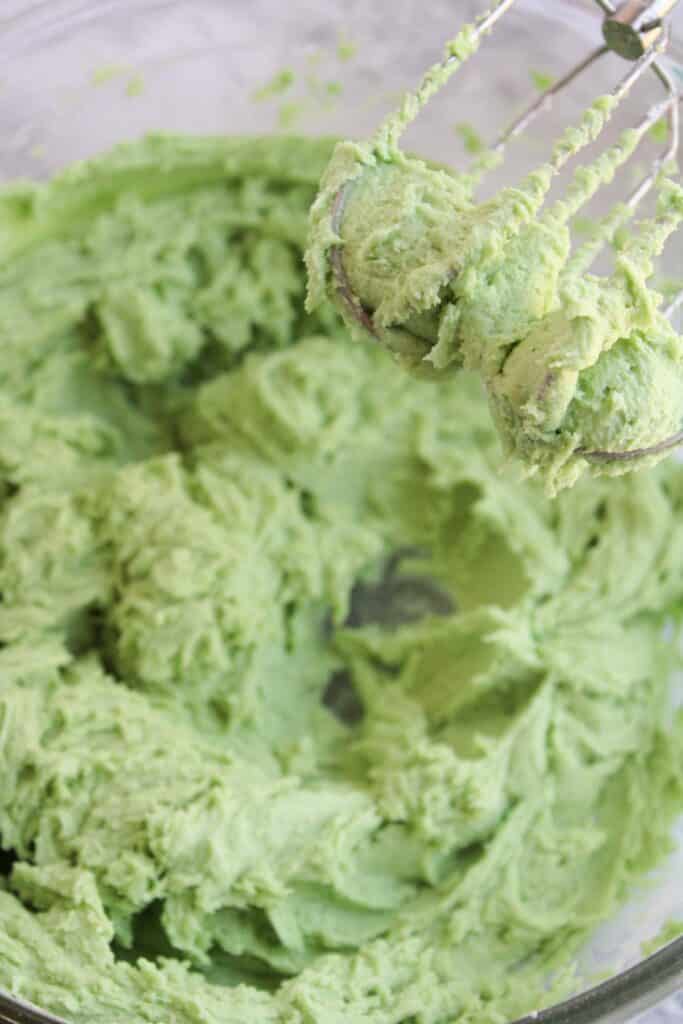 A close-up of green cookie dough in a bowl with a hand mixer featuring beaters coated in the dough, perfect for making Festive Christmas Tree Cupcakes.