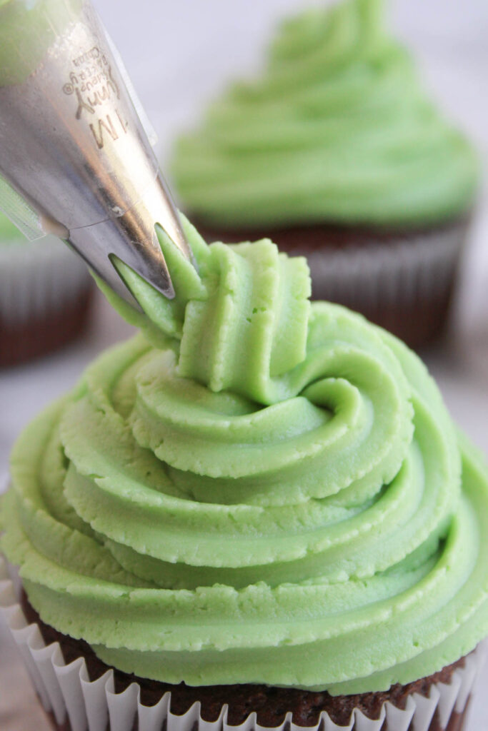 A piping bag with a star nozzle is applying green frosting onto a cupcake, creating the look of festive Christmas tree cupcakes. Another frosted cupcake is in the background.