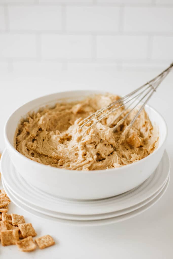 A white mixing bowl filled with a light brown, creamy batter sits on a stack of glazed cinnamon plates. A whisk is resting inside the bowl, and some cereal pieces reminiscent of crunch toast donuts are scattered around the base.