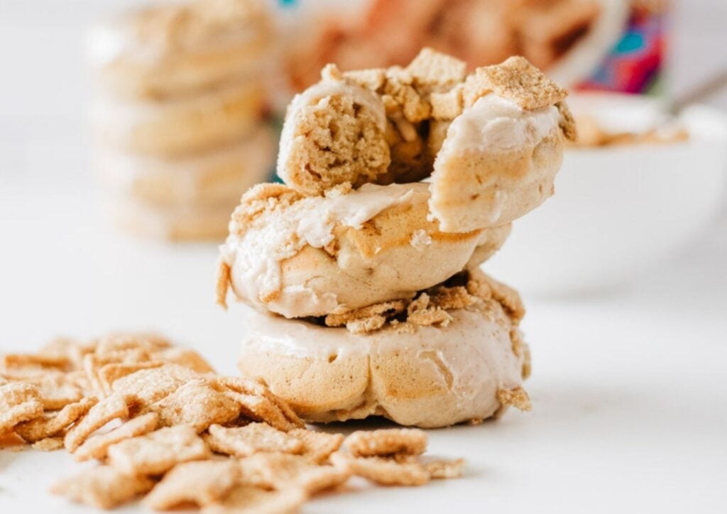 Three frosted donuts stacked on a white surface, topped and surrounded by pieces of Glazed Cinnamon Crunch Toast donuts.
