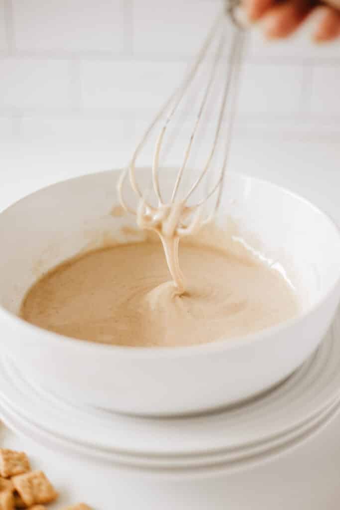 A whisking hand stirs a creamy, beige mixture in a white bowl on a white surface, prepping for some delectable glazed cinnamon crunch toast donuts.