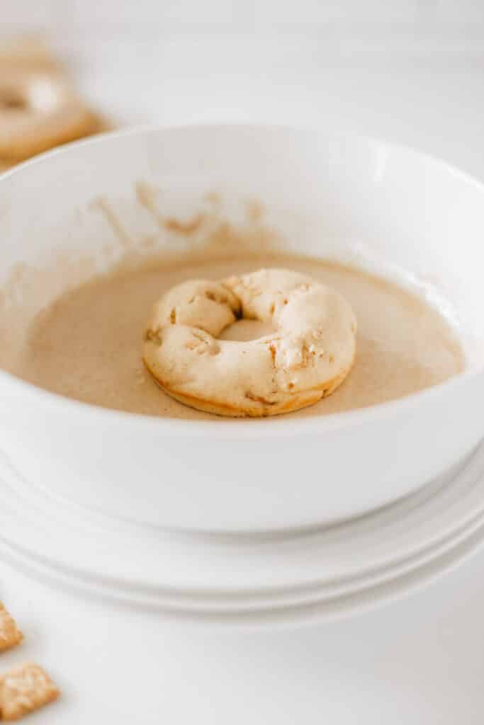 A glazed donut is partially submerged in a bowl of batter, surrounded by a few pieces of cinnamon crunch toast on a white surface.