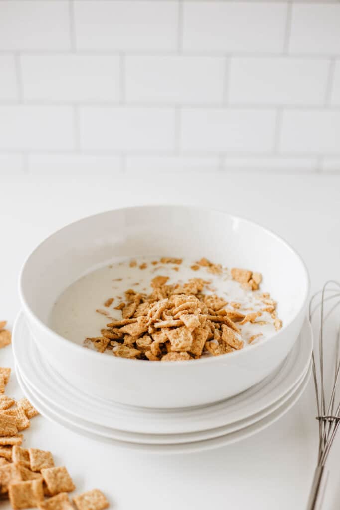 A white bowl filled with milk and cereal sits on stacked plates. Some pieces of cereal are scattered on the white surface, mingling with a couple of glazed cinnamon crunch toast donuts. A wire whisk is placed next to the bowl.