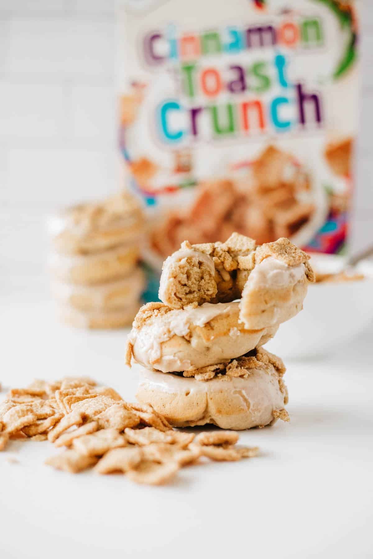 Three glazed Cinnamon Toast Crunch donuts are stacked in front of a Cinnamon Toast Crunch cereal box, with cereal pieces scattered around and additional donuts in the background.