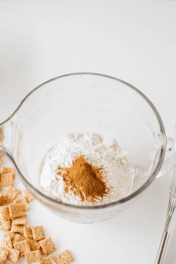 A glass mixing bowl contains flour and a pile of cinnamon on top, hinting at the creation of glazed cinnamon crunch toast donuts. Nearby, there are pieces of cereal and a whisk on a pristine white surface.