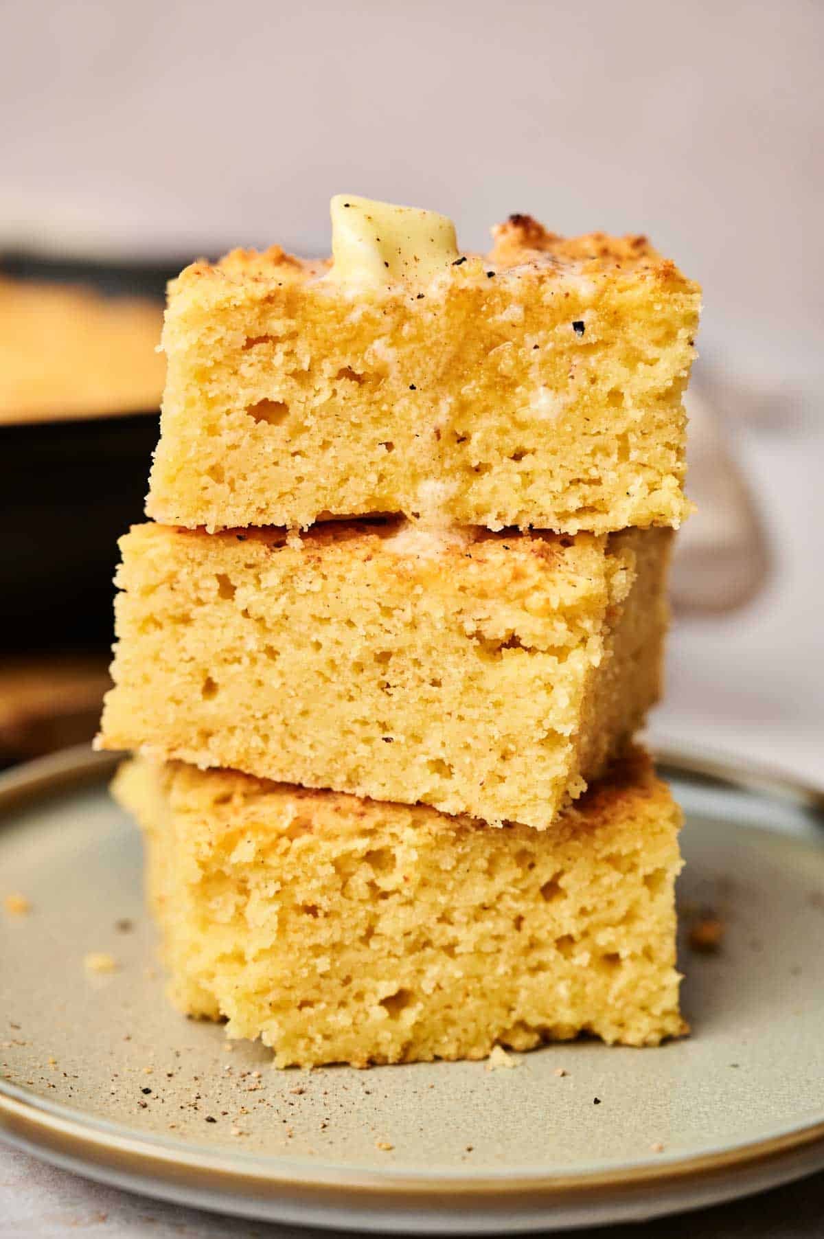 A stack of three moist cornbread pieces on a plate with a small piece of butter melting on top.