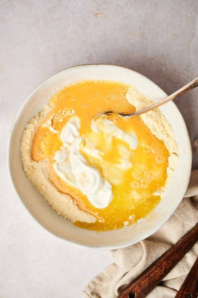 A mixing bowl containing flour, eggs, and cream, with a spoon partially submerged in the mixture, hints at the beginnings of cornbread. A cloth and wooden utensils are placed beside the bowl.
