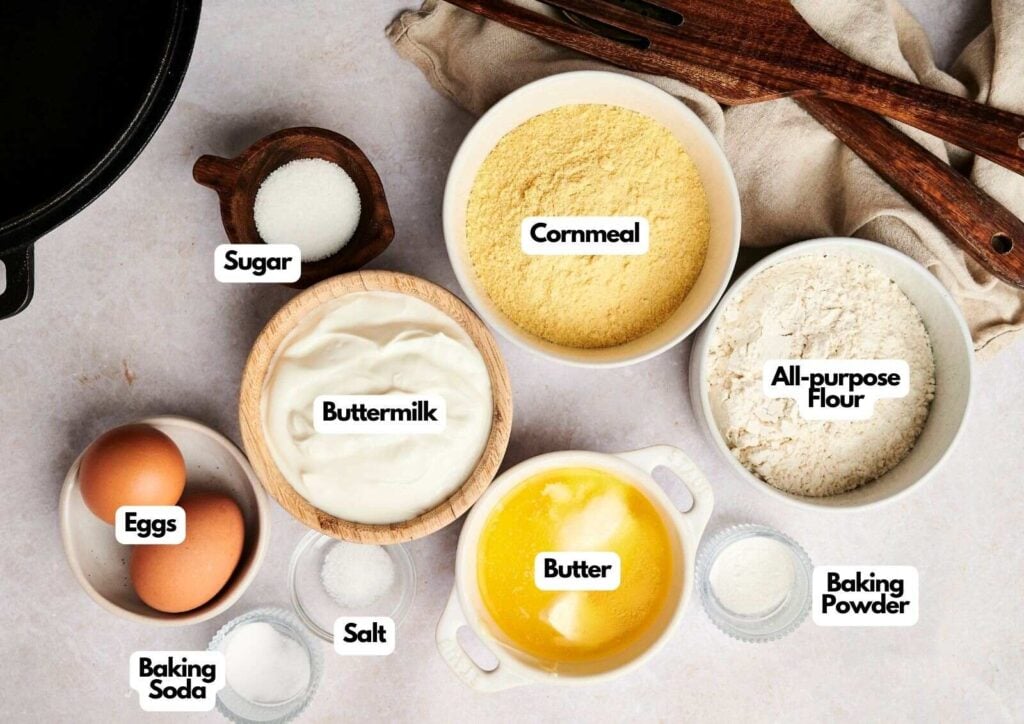 Ingredients for classic cornbread, including cornmeal, flour, buttermilk, eggs, butter, sugar, salt, baking powder, and baking soda, are arranged in bowls on a light surface.