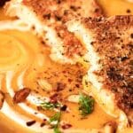 A bowl of creamy butternut squash soup with herb-crusted toast pieces on top, garnished with seeds and herbs.
