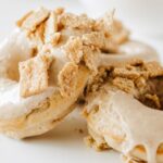 Two doughnuts with a light-colored glaze are topped with crumbled cinnamon crunch toast, placed on a white surface.