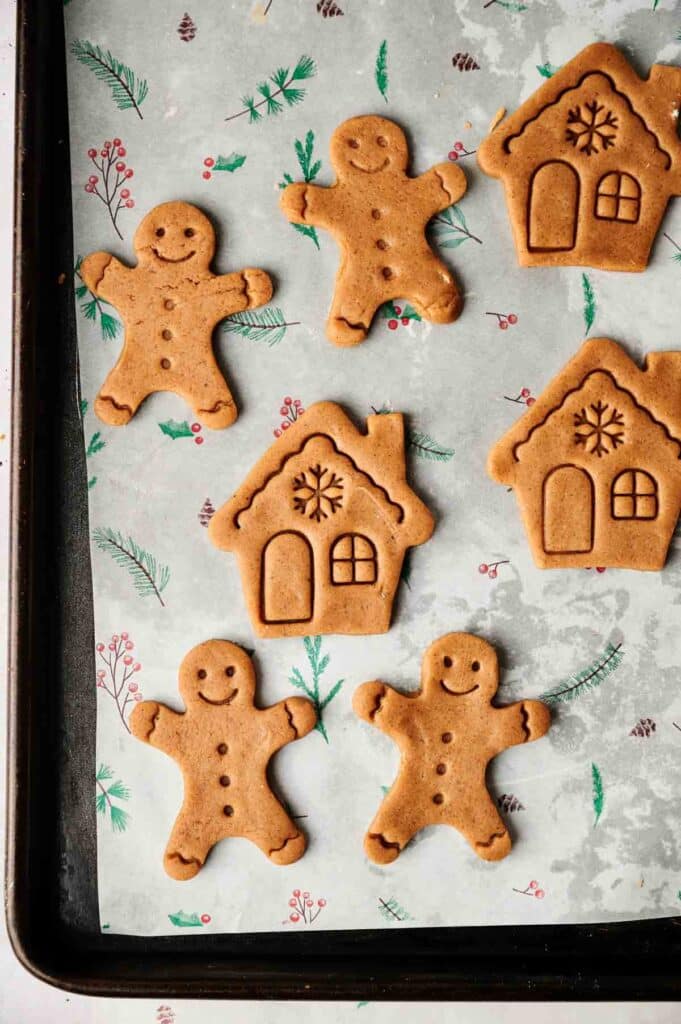 Gingerbread cookies shaped like houses and gingerbread men rest on a parchment-lined baking sheet, adorned with festive holiday-themed decorations.