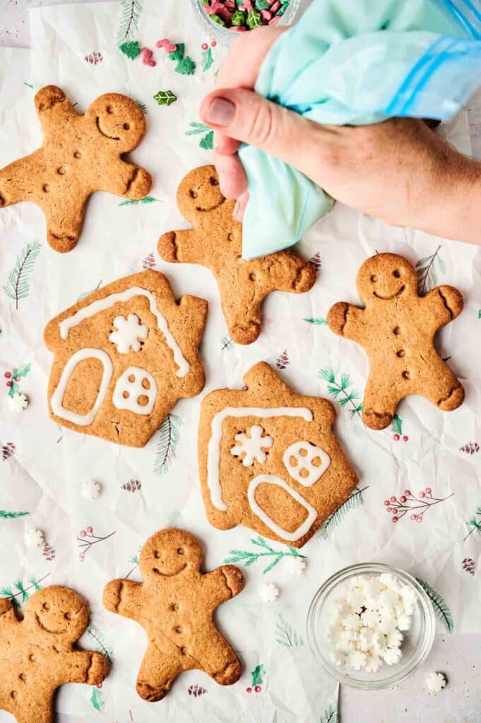 A hand decorates Gingerbread Cookies with white icing. The cookies are shaped like gingerbread people and houses, arranged on festive paper alongside a container of decorative sprinkles.