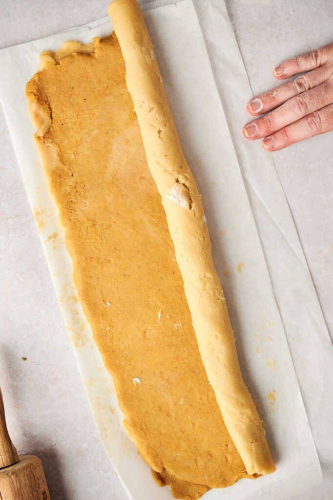 Someone is rolling dough on parchment paper on a countertop, their fingers slightly covered in flour. A rolling pin, ready to shape the Pumpkin Spice Roll Cookies, is partially visible in the lower left corner.