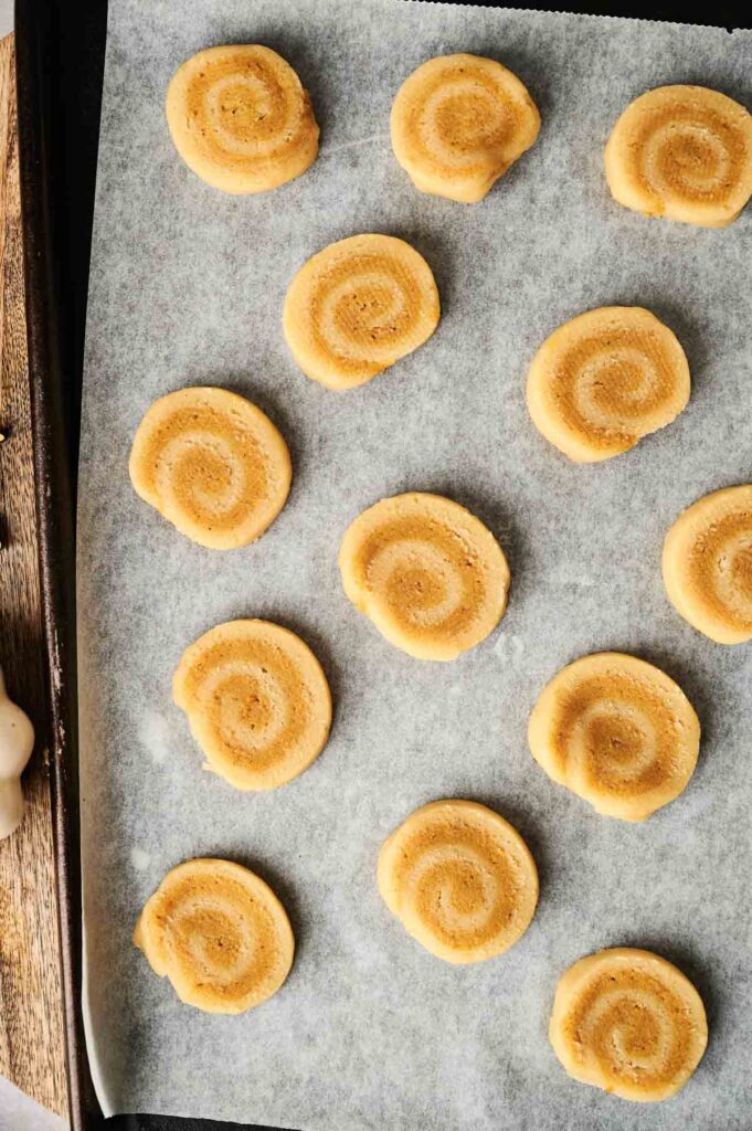 A baking sheet lined with parchment paper holds 15 evenly spaced, spiral-shaped Pumpkin Spice Roll Cookies ready for baking.