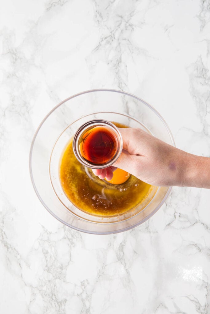 A hand holds a small glass bowl of vanilla extract above a mixing bowl containing eggs and brown liquid on a marble countertop, preparing the base for Raspberry and White Chocolate Blondies.