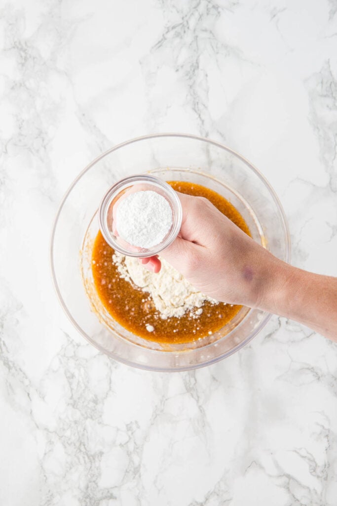 A hand holds a small bowl of white powder over a glass bowl containing flour and a brown liquid mixture on a marble surface, preparing ingredients for delicious Raspberry and White Chocolate Blondies.