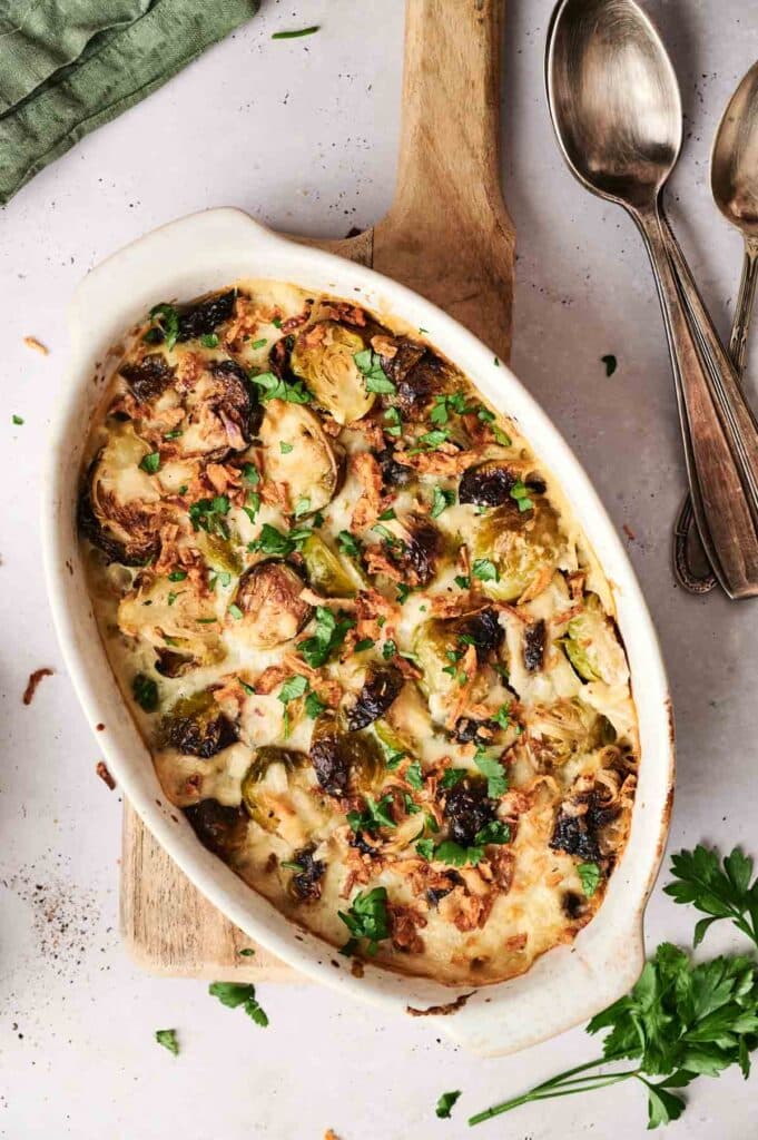 A baked casserole dish filled with browned Brussels sprouts and garnished with fresh parsley. Spoons are placed beside the dish on a wooden board.