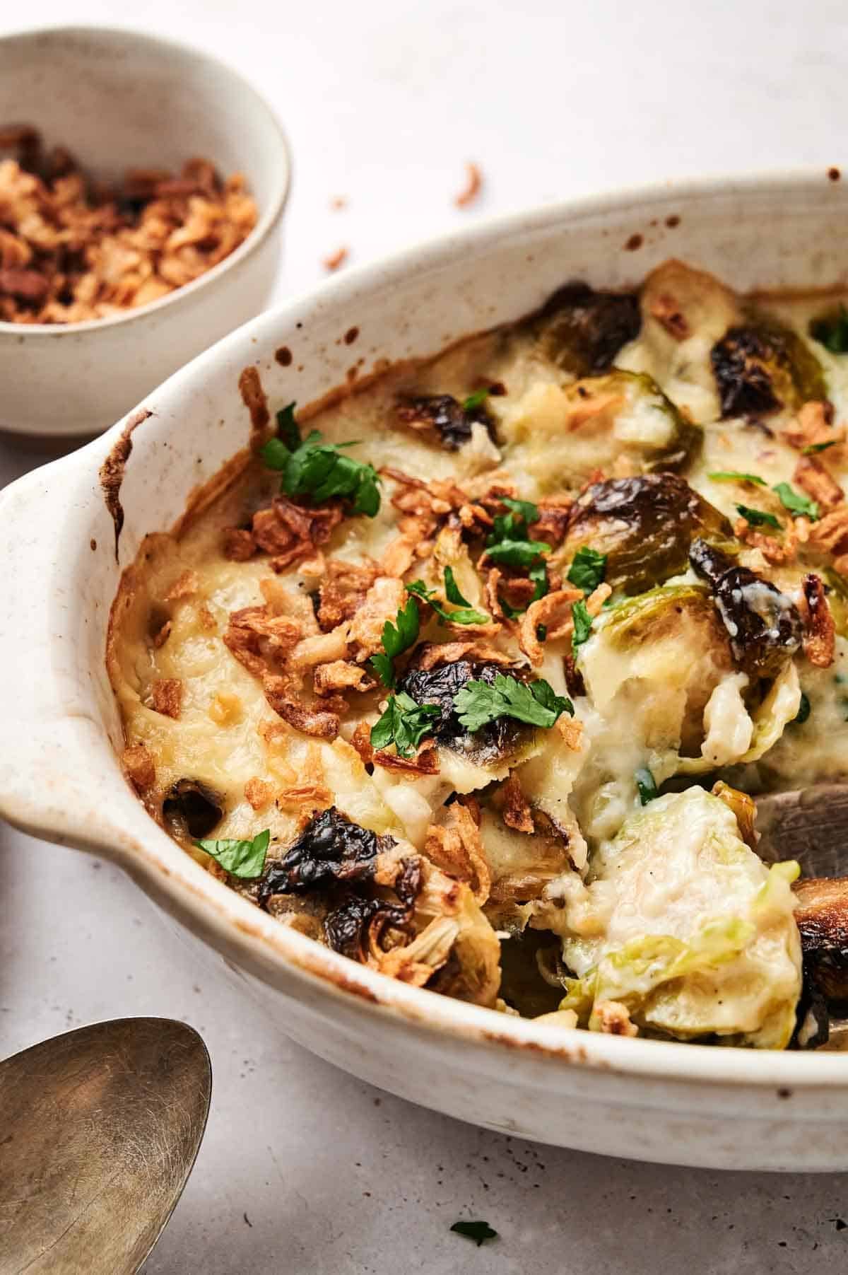 A baked dish with melted cheese on top, garnished with herbs and crispy onions, in a white ceramic dish. A serving spoon is nearby, with a small bowl of extra toppings in the background.