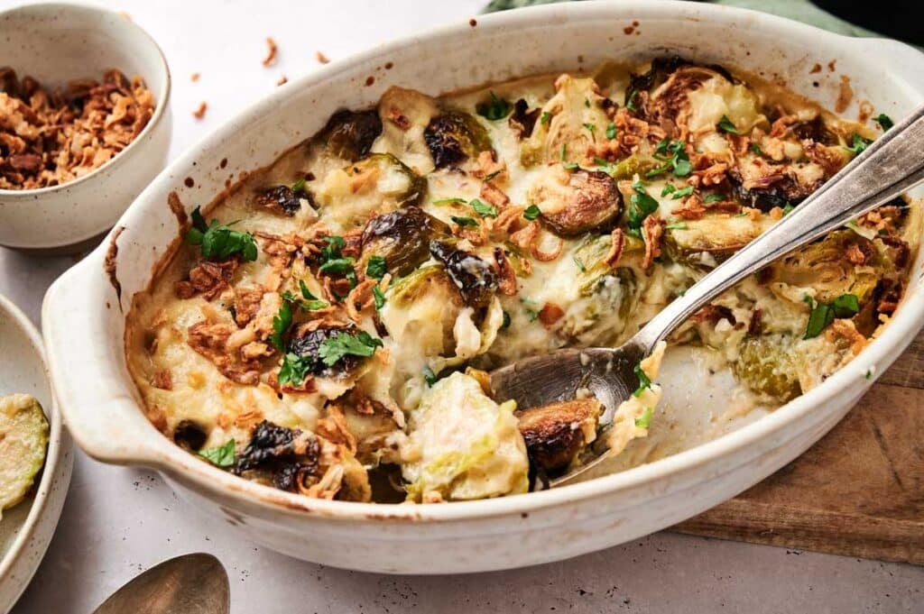 A baked dish with Brussels sprouts, topped with herbs and crispy bits, placed in an oval-shaped ceramic dish with a serving spoon.