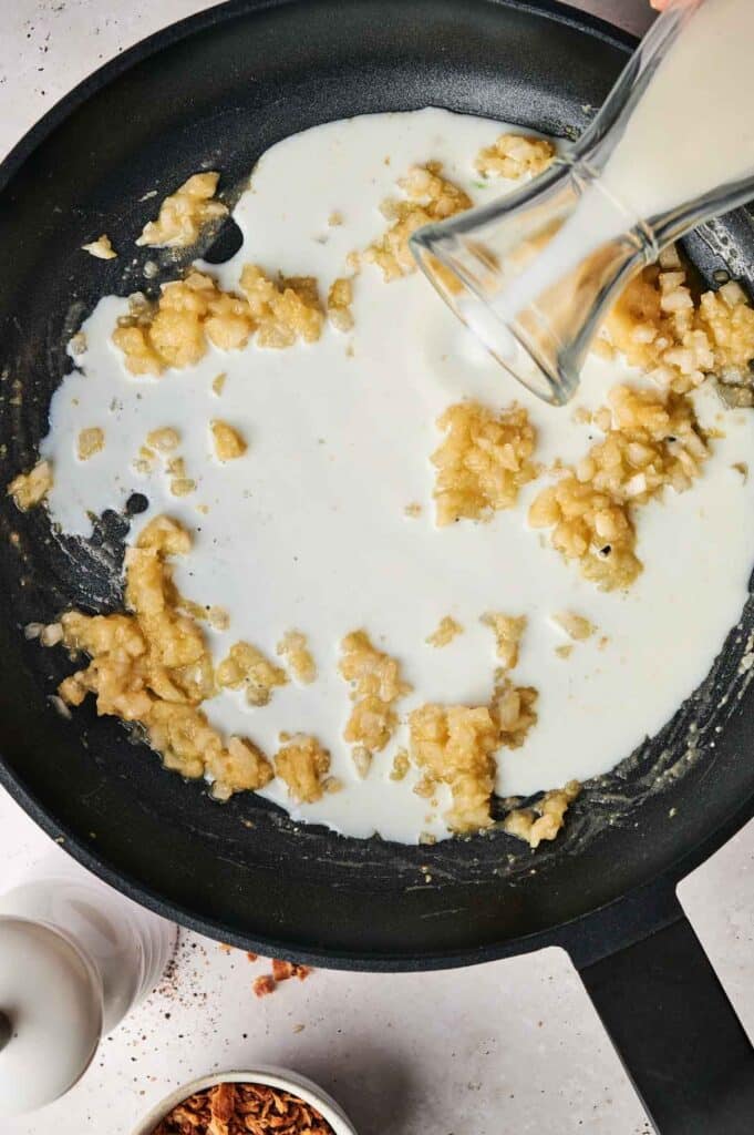 Milk being poured into a black frying pan containing mashed garlic and butter, with a spice jar nearby.