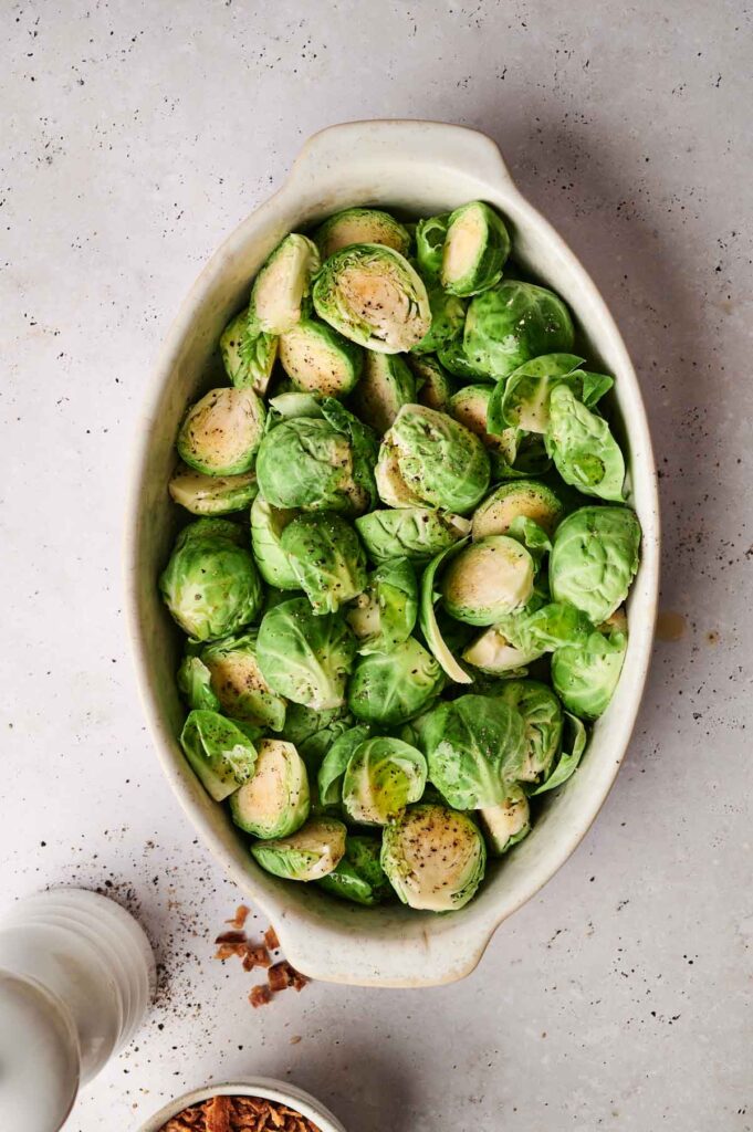 Top view of a ceramic dish filled with halved Brussels sprouts, seasoned with pepper. A white pepper mill and a small bowl with crushed nuts are nearby on a light surface.