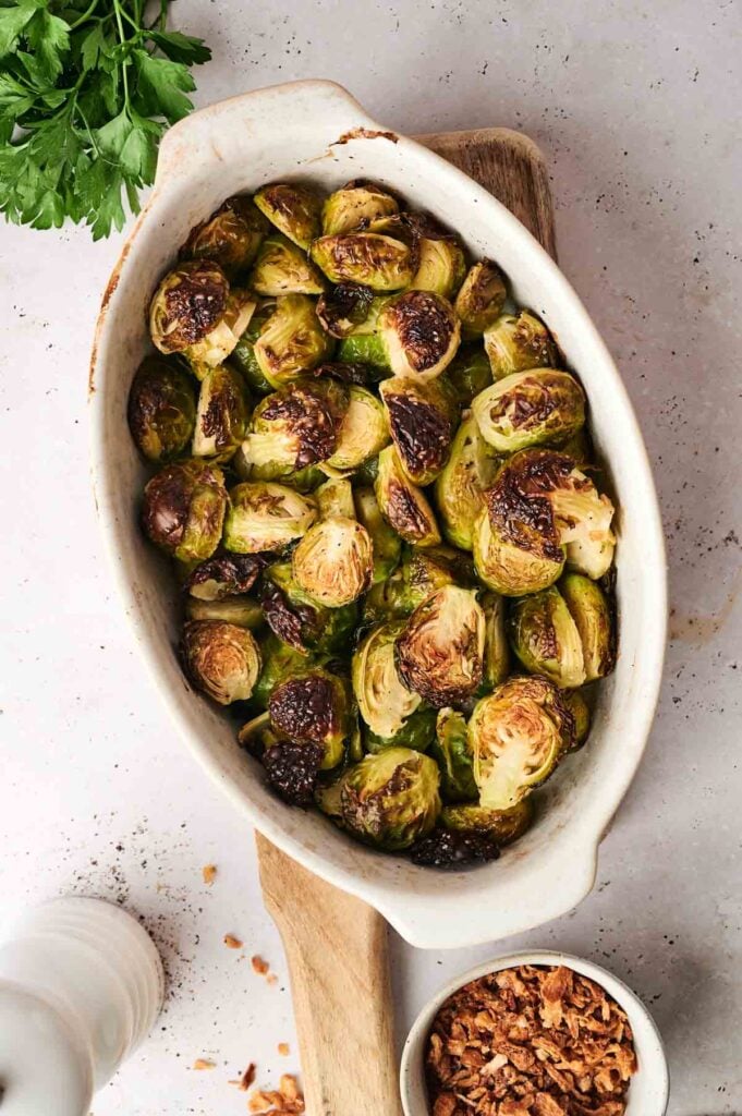 Roasted Brussels sprouts in an oval dish on a wooden board, with chopped parsley and a small bowl of crispy onions on the side.