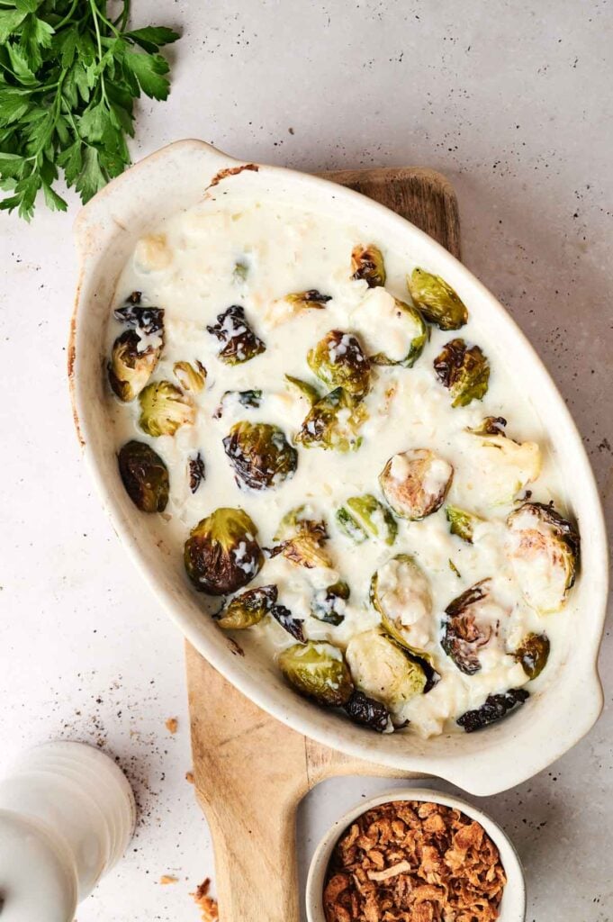 A casserole dish with creamy Brussels sprouts and a bowl of crispy fried onions on a wooden board, garnished with fresh parsley.