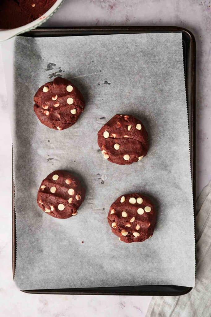 Four Copycat Crumbl Red Velvet White Chip Cookies rest invitingly on a parchment-lined baking tray.