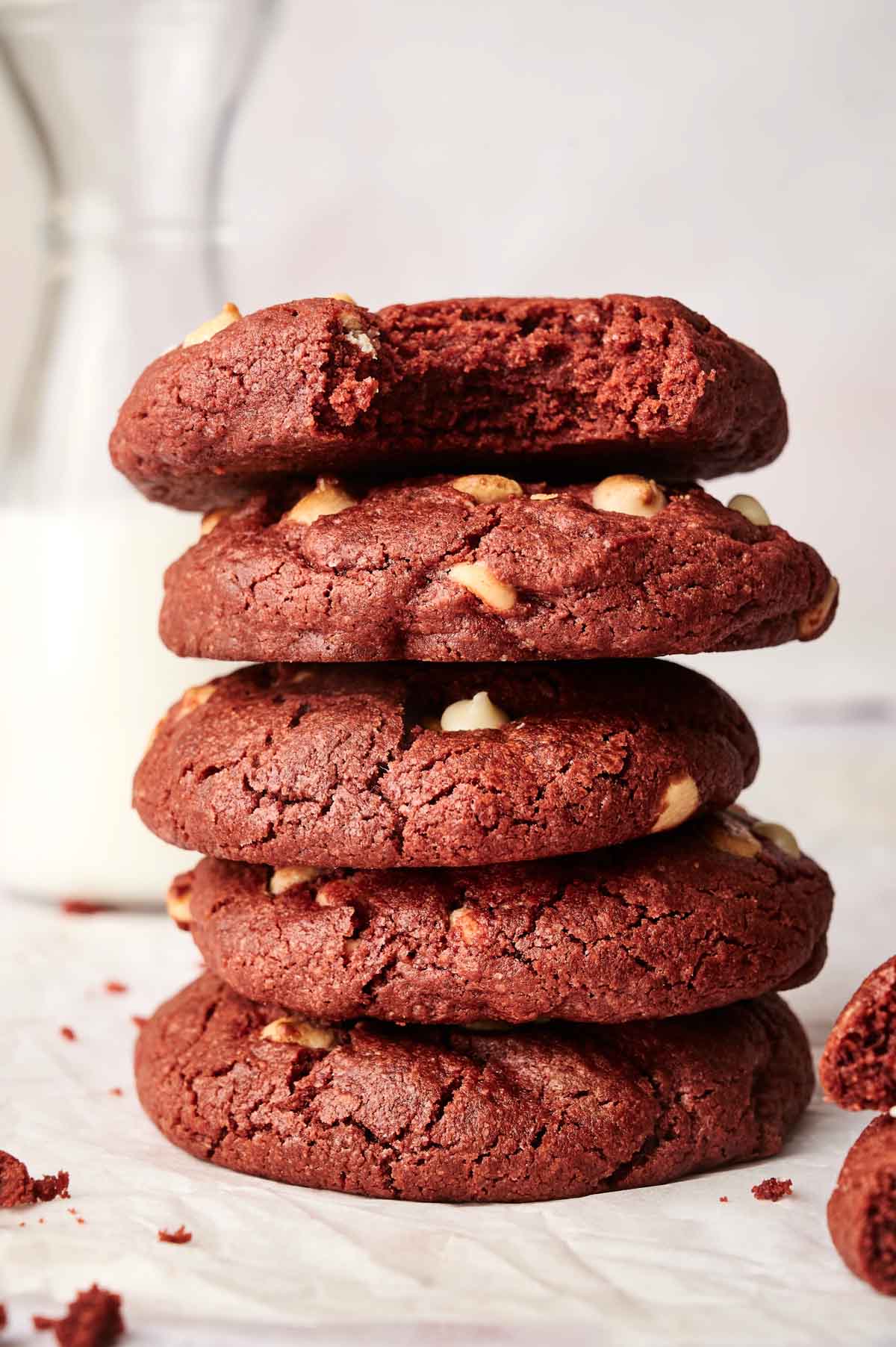 A stack of five Copycat Crumbl Red Velvet White Chip Cookies with white chocolate chips stands tall. A glass of milk awaits in the background, ready to accompany this delightful treat.