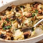 A baked Brussels sprouts dish with creamy sauce, topped with crispy onions and parsley, shown in a white oval dish with a spoon.