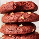 A stack of three copycat Crumbl red velvet cookies with delightful white chocolate chips sits invitingly. The top cookie, with a tempting bite missing, promises a delicious treat.