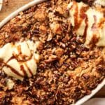 A baked dessert with a crumbly pecan topping, drizzled with sauce, and topped with scoops of melting ice cream in an oval dish on a wooden surface, this Pumpkin Dump Cake delights the senses with its perfect blend of flavors.