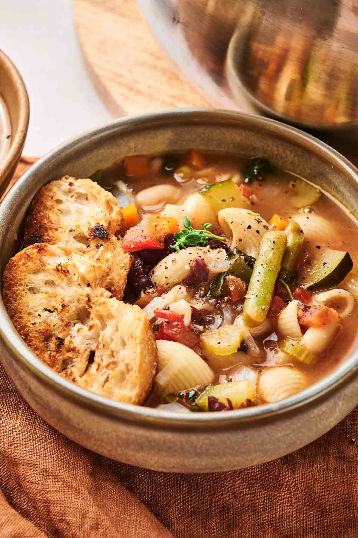 A bowl of minestrone soup with pasta and toasted bread slices on the side.