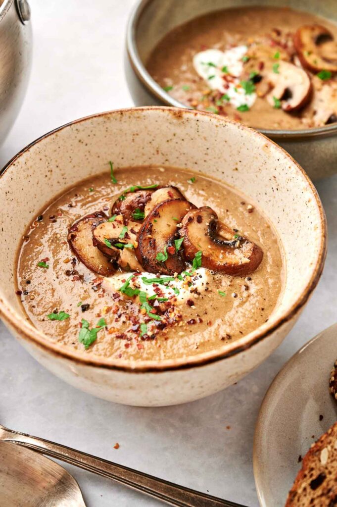 A bowl of delectable mushroom soup, garnished with sliced mushrooms, aromatic herbs, and a sprinkle of red pepper flakes, sits invitingly next to a shining spoon.