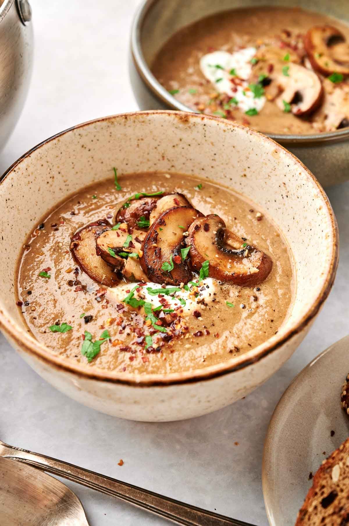 A bowl of delectable mushroom soup, garnished with sliced mushrooms, aromatic herbs, and a sprinkle of red pepper flakes, sits invitingly next to a shining spoon.