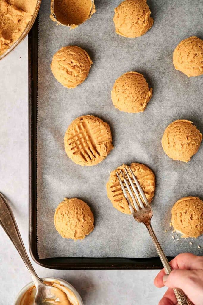 Close-up of peanut butter cookie dough scoops on a baking sheet lined with parchment paper. A hand presses a fork onto a dough ball, creating the signature crisscross pattern.