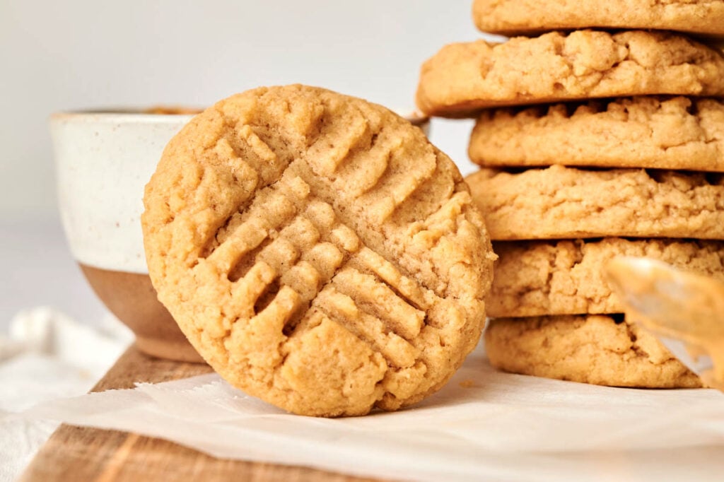 A close-up of a classic Peanut Butter Cookies with a crisscross fork pattern, leaning against a stack of similar cookies on a wooden surface.