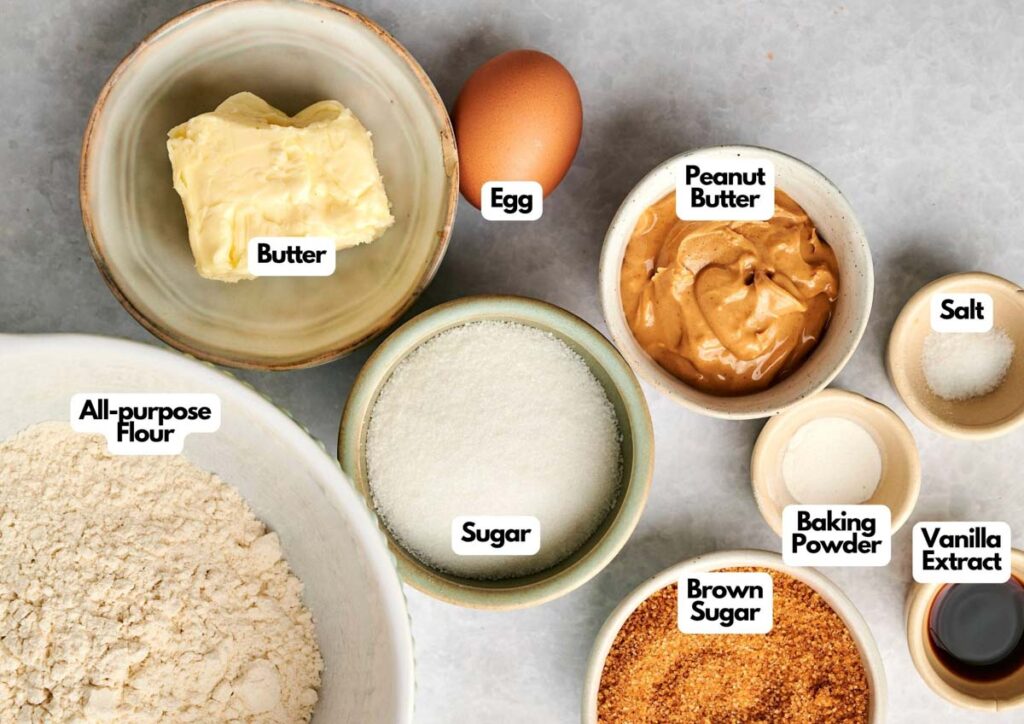 Top view of baking ingredients for Peanut Butter Cookies in bowls, including butter, egg, peanut butter, all-purpose flour, sugar, brown sugar, baking powder, vanilla extract, and salt—all clearly labeled.